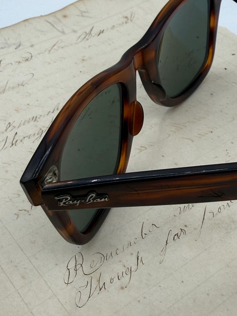 May include: Close-up of Ray-Ban sunglasses with a tortoiseshell frame. The sunglasses have dark green lenses and the Ray-Ban logo is visible on the arm. The glasses are resting on a piece of paper with handwritten script.