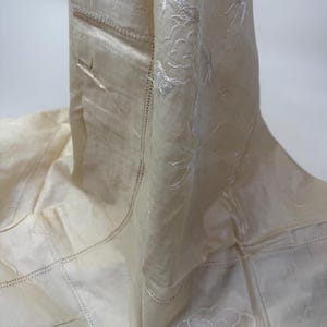 May include: Off-white fabric with embroidered floral and cloud designs. The fabric has a stitched border and a patchwork design. The embroidery is in a lighter shade of off-white, creating a subtle contrast. The fabric is draped, showing its texture and design.