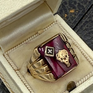 May include: A gold-toned ring with a rectangular, deep red stone. The ring features a small diamond-like accent and a gold emblem. It is presented in a cream-colored ring box with a satin interior.