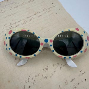 May include: Oval-shaped sunglasses with a cream-colored frame decorated with pink and blue polka dots. The lenses are black, and the arms are white. The sunglasses are resting on a piece of aged paper with handwritten text.