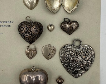 May include: A collection of silver heart-shaped pendants and lockets. The pendants vary in size and design, including smooth, engraved, and openable styles. Some have intricate floral patterns, while others are plain.
