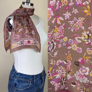 May include: A brown rectangular scarf featuring a floral pattern in shades of pink, yellow, and white. The scarf has a decorative border with a Greek key geometric design and circular motifs, finished with a thin hem along the edges.