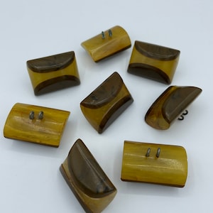 May include: Eight brown and gold plastic buttons with two holes each. The buttons have a rounded rectangular shape with a curved top edge.