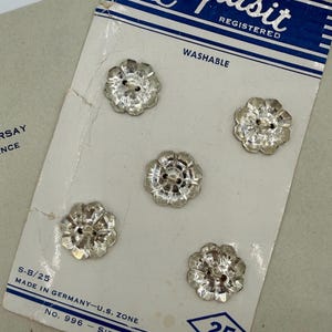 May include: A card of five clear, flower-shaped buttons. The buttons are made of a shiny, possibly glass or crystal material. The card reads "Exquisit" and "Washable". The card also states "Made in Germany-U.S. Zone".