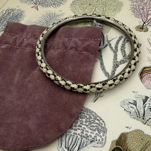 May include: A silver-toned bracelet with a woven cream and white pattern. The bracelet is next to a small, dark purple velvet pouch. The background features a vintage botanical illustration with coral and plant drawings.