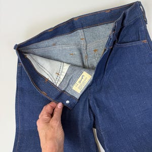 Puede incluir: Primer plano de unos vaqueros de mezclilla azul oscuro, desabrochados para mostrar el forro interior y una etiqueta blanca de la marca "Wrangler". Las costuras naranjas contrastan con la mezclilla. Los vaqueros son de material resistente.