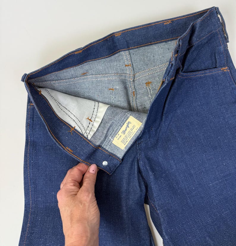 Puede incluir: Primer plano de unos vaqueros de mezclilla azul oscuro, desabrochados para mostrar el forro interior y una etiqueta blanca de la marca "Wrangler". Las costuras naranjas contrastan con la mezclilla. Los vaqueros son de material resistente.