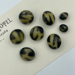 May include: Eight vintage buttons with a marbled design in olive green and black. The buttons are of varying sizes and are arranged on a cream-coloured paper. The paper has text that reads "OTEL" and "LEANS 12".
