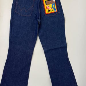 Pode incluir: Jeans Wrangler de ganga azul escuro com perna larga. Os jeans t&ecirc;m um design cl&aacute;ssico de cinco bolsos com costuras laranja. Uma etiqueta Wrangler est&aacute; presa ao bolso traseiro e o tamanho &eacute; 28x29.