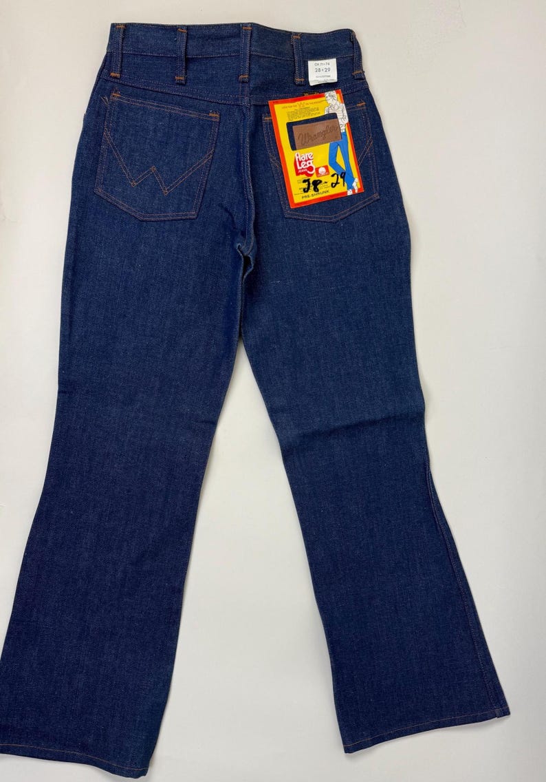 Pode incluir: Jeans Wrangler de ganga azul escuro com perna larga. Os jeans t&ecirc;m um design cl&aacute;ssico de cinco bolsos com costuras laranja. Uma etiqueta Wrangler est&aacute; presa ao bolso traseiro e o tamanho &eacute; 28x29.