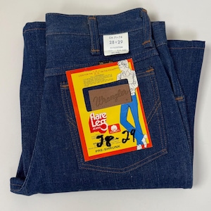 Pode incluir: Jeans Wrangler flare azul escuro, tamanho 28x29, com etiqueta amarela e vermelha. A etiqueta apresenta um desenho de uma pessoa usando jeans. Os jeans s&atilde;o pr&eacute;-encolhidos e de algod&atilde;o.