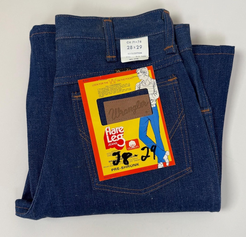 Pode incluir: Jeans Wrangler flare azul escuro, tamanho 28x29, com etiqueta amarela e vermelha. A etiqueta apresenta um desenho de uma pessoa usando jeans. Os jeans s&atilde;o pr&eacute;-encolhidos e de algod&atilde;o.