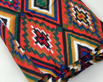 May include: A folded fabric with a geometric pattern in shades of red, orange, yellow, green, blue, black, and white. The pattern features repeating diamond shapes with intricate details.