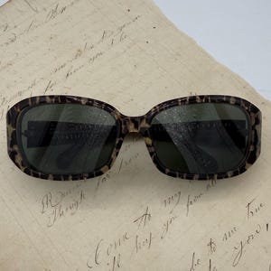 Può includere: Occhiali da sole quadrati con motivo tartarugato. Le lenti sono scure e la montatura ha un design maculato marrone e beige. Gli occhiali da sole sono appoggiati su un pezzo di carta invecchiata con scritte a mano.