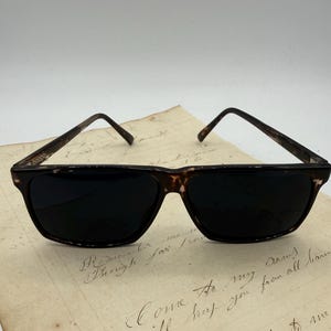 Può includere: Occhiali da sole con montatura a fantasia tartarugata e lenti scure. Le montature rettangolari hanno un design classico. Gli occhiali da sole sono appoggiati su uno sfondo di carta vintage.