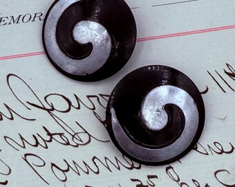 Könnte beinhalten: Zwei runde, schwarze Knöpfe mit einem silbernen, spiralförmigen Design. Die Knöpfe sind vor einem Hintergrund aus handschriftlichem Text auf gealtertem Papier platziert.