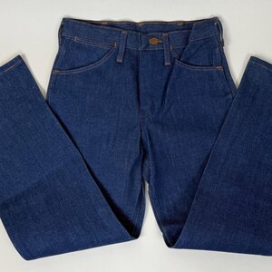 Puede incluir: Vaqueros de mezclilla azul oscuro con un dise&ntilde;o cl&aacute;sico de cinco bolsillos y una pernera acampanada. Los vaqueros tienen cierre de bot&oacute;n y cremallera, y las costuras est&aacute;n cosidas con hilo naranja en contraste. Los vaqueros est&aacute;n planos sobre una superficie blanca.
