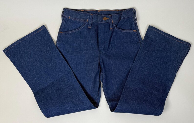 Puede incluir: Vaqueros de mezclilla azul oscuro con un dise&ntilde;o cl&aacute;sico de cinco bolsillos y una pernera acampanada. Los vaqueros tienen cierre de bot&oacute;n y cremallera, y las costuras est&aacute;n cosidas con hilo naranja en contraste. Los vaqueros est&aacute;n planos sobre una superficie blanca.