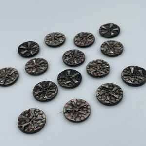 May include: A collection of 14 round, black and silver buttons with a starburst design. The buttons have a textured surface and a raised, silver starburst design in the center.