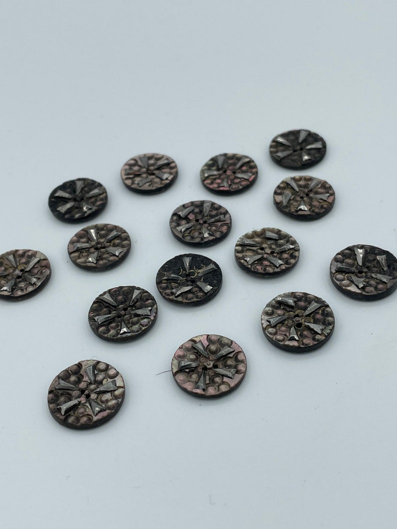 May include: A collection of 14 round, black and silver buttons with a starburst design. The buttons have a textured surface and a raised, silver starburst design in the center.