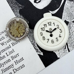 May include: Two vintage clock-themed buttons. One button has a silver-colored, textured frame with clear rhinestones and a gold-colored clock face. The other is a white, round button with a clock face and black hands.