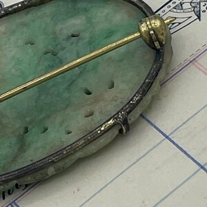 May include: A green jade oval brooch with a silver setting and a gold pin. The brooch has a carved design and is set on a white background.