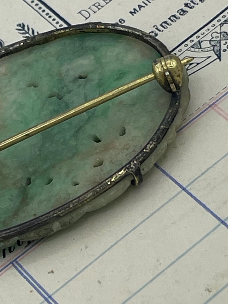 May include: A green jade oval brooch with a silver setting and a gold pin. The brooch has a carved design and is set on a white background.