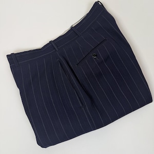 May include: Navy blue pinstripe trousers. The pants have a classic design with vertical stripes and a buttoned back pocket. The trousers are folded and laid flat on a white surface.