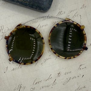 May include: Vintage round sunglasses with a tortoiseshell pattern frame and dark lenses. The glasses are connected by a thin silver wire and rest on a handwritten letter. A black leather case is in the background.