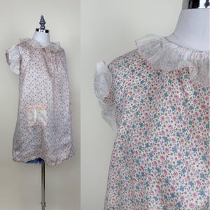 May include: A vintage floral print dress with a white ruffled collar and sleeves. The dress features a small pocket and is displayed on a mannequin. The fabric has a pattern of small blue, pink, and green flowers on a white background.