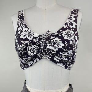 May include: A sleeveless crop top featuring a black and white floral pattern. The garment has a scoop neckline and a central ruched detail with a small tie at the center of the chest, creating a gathered effect across the front of the bodice.