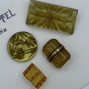 May include: A collection of vintage buttons in shades of amber and olive green. The buttons feature various designs, including floral patterns and geometric shapes. One button has a decorative metal band. The buttons are arranged on a white surface.