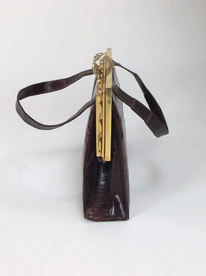 May include: A brown leather handbag with a gold clasp and a chain strap. The bag has a rectangular shape and is made of a textured leather.