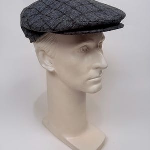May include: A grey flat cap featuring a dark grey diamond grid pattern. The cap has a structured crown and a short, stiff brim that extends from the front. The fabric has a textured appearance, and the design covers the entire top surface.