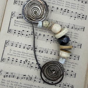 May include: A handcrafted metal bookmark with a spiral design at each end, connected by a slender wire. The bookmark features a series of beads in various shapes and colors, including black, white, and beige, resting on a vintage sheet music background.
