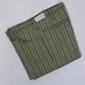 Puede incluir: Pantalones verde oliva doblados con rayas verticales azules y rojas. Los pantalones tienen un diseño clásico con cierre de botón y cremallera, trabillas para el cinturón y bolsillos. Una pequeña etiqueta está adherida a la cintura.