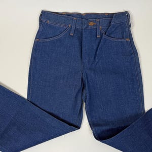 Puede incluir: Vaqueros de mezclilla azul oscuro con un dise&ntilde;o cl&aacute;sico de cinco bolsillos. Los vaqueros tienen cierre de bot&oacute;n y corte recto. Las costuras son de color naranja en contraste. Los vaqueros est&aacute;n colocados sobre una superficie blanca.