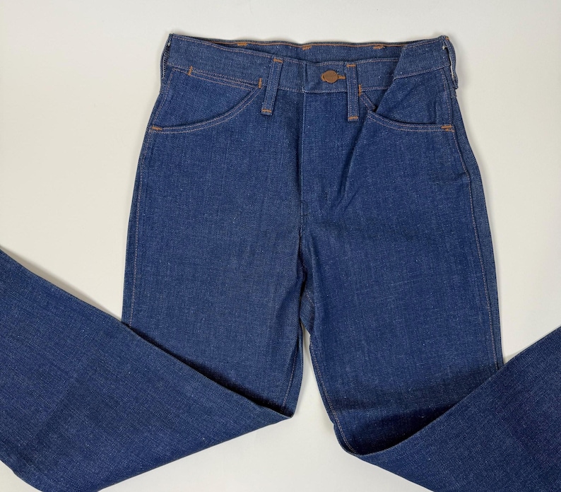 Puede incluir: Vaqueros de mezclilla azul oscuro con un dise&ntilde;o cl&aacute;sico de cinco bolsillos. Los vaqueros tienen cierre de bot&oacute;n y corte recto. Las costuras son de color naranja en contraste. Los vaqueros est&aacute;n colocados sobre una superficie blanca.