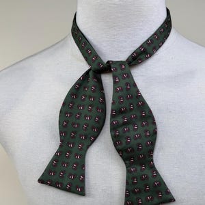 Puede incluir: Pajarita verde oscuro con un estampado de cachemira repetido en burdeos y blanco. La pajarita se muestra en un maniquí blanco, mostrando su forma y diseño. La corbata está anudada al cuello.