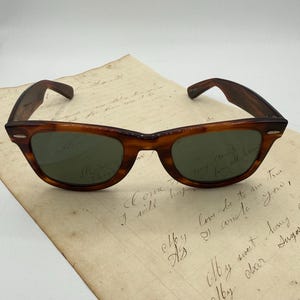 May include: Classic tortoiseshell sunglasses with green lenses sit on a handwritten letter. The sunglasses have a vintage design with a curved browline and thick arms. The letter features cursive handwriting with faded ink.