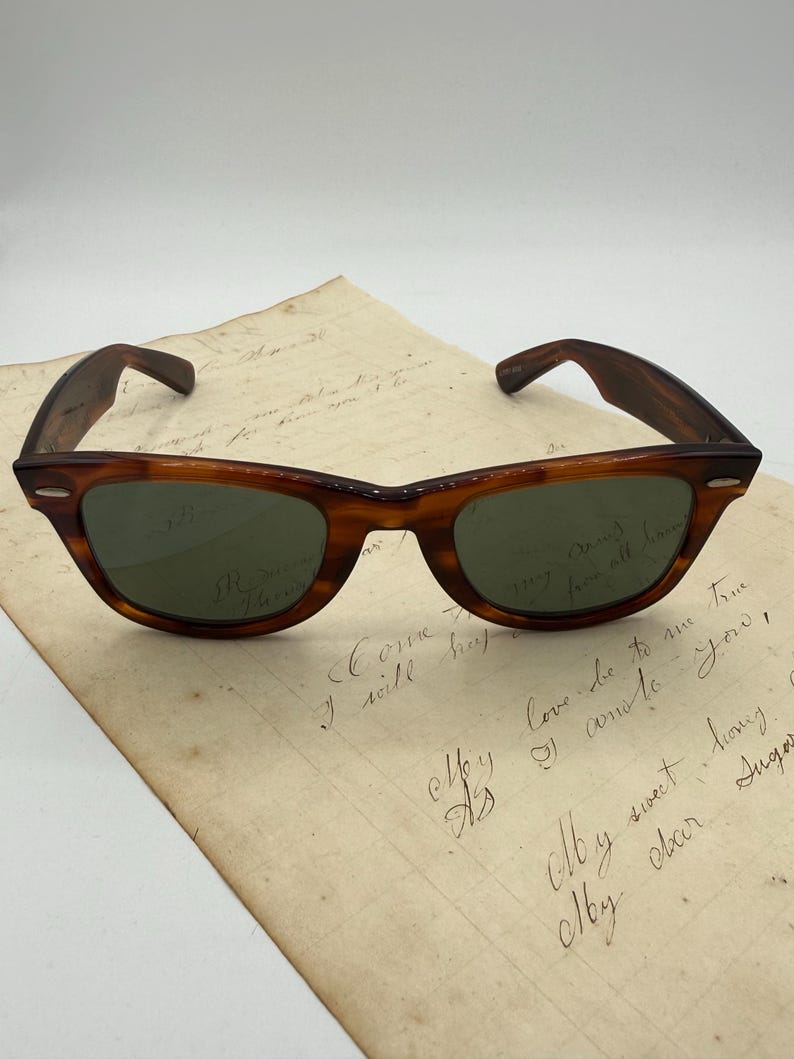 May include: Classic tortoiseshell sunglasses with green lenses sit on a handwritten letter. The sunglasses have a vintage design with a curved browline and thick arms. The letter features cursive handwriting with faded ink.