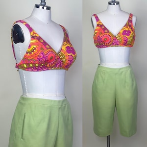 May include: A vintage two-piece set featuring a colorful floral print bikini top in shades of pink, orange, and yellow. Paired with high-waisted, light green shorts. The set is displayed on a mannequin.