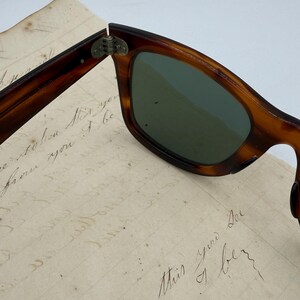 May include: Close-up of vintage sunglasses with a tortoiseshell frame and green lenses. The sunglasses are partially open, revealing the hinge. The background includes a piece of aged paper with handwritten script.