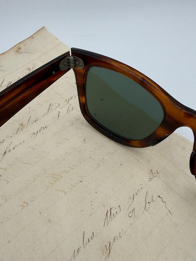 May include: Close-up of vintage sunglasses with a tortoiseshell frame and green lenses. The sunglasses are partially open, revealing the hinge. The background includes a piece of aged paper with handwritten script.