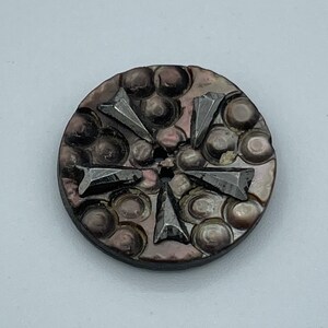 May include: A round, black and iridescent button with a raised, geometric design. The button has a hole in the center and is made of mother of pearl.