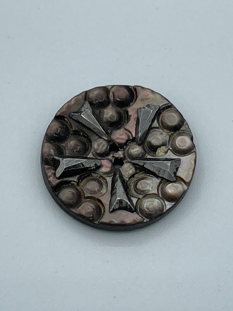 May include: A round, black and iridescent button with a raised, geometric design. The button has a hole in the center and is made of mother of pearl.
