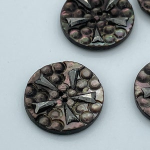 May include: A set of six iridescent black shell buttons with a raised starburst design. The buttons have a smooth, shiny finish and a hole in the center for sewing.