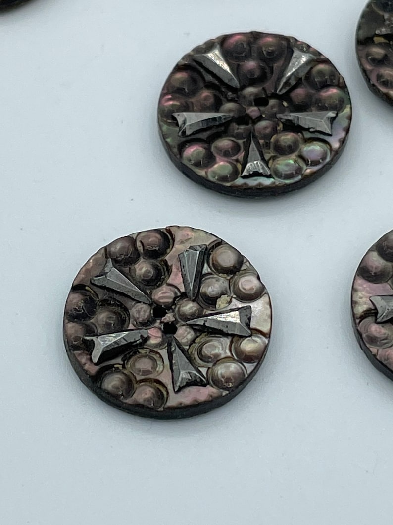 May include: A set of six iridescent black shell buttons with a raised starburst design. The buttons have a smooth, shiny finish and a hole in the center for sewing.