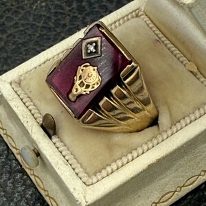 Könnte beinhalten: Ein goldfarbener Ring mit einem rechteckigen, tiefroten Stein. Der Ring hat einen kleinen Diamanten und ein goldenes Emblem. Er wird in einer cremefarbenen Schmuckschatulle mit goldenem Rand präsentiert.