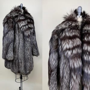Puede incluir: Abrigo largo de piel de zorro plateado exhibido en un maniquí. El abrigo presenta una textura rica y densa, con una mezcla de tonos oscuros y plateados. El pelaje es denso y suave, con un cuello ancho. Una prenda de abrigo llamativa.